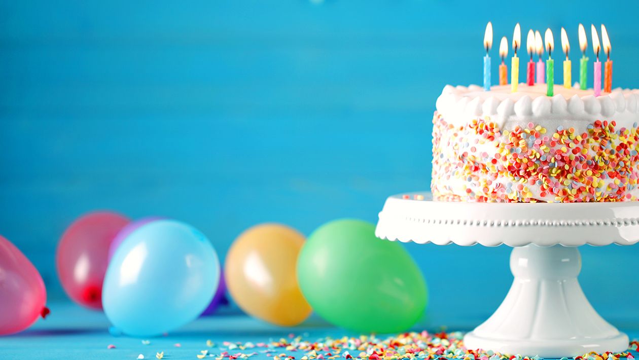Delicious birthday cake on a white tray with burning candles and colorful balloons on table